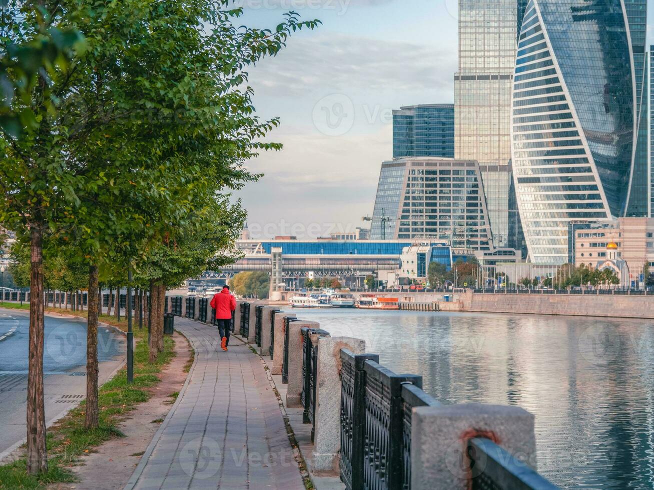 Morgen Fitness Lauf auf das Damm im Moskau. ein Mann im ein rot Sport Windjacke läuft Nieder ein verlassen Straße früh im das Morgen foto