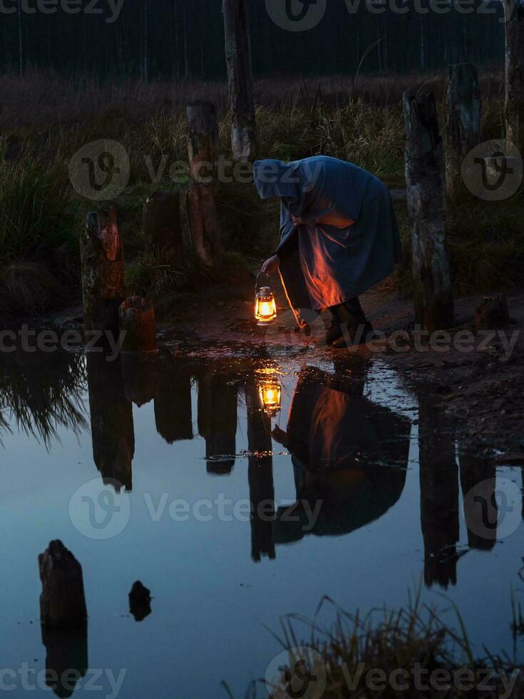 gebogen Mann halten das alt Lampe draußen in der Nähe von das See. Hand hält ein groß Lampe im das dunkel. uralt Kerosin Laterne leuchtet das Weg auf ein Nacht. Licht und hoffen Konzept. foto