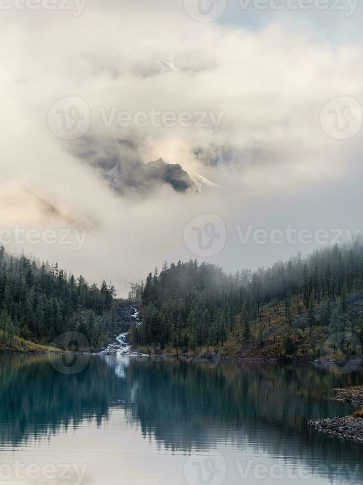 atmosphärisch alpin Landschaft mit Bäume in der Nähe von Berg See und hoch schneebedeckt Berg im niedrig Wolken. verisch Sicht. foto