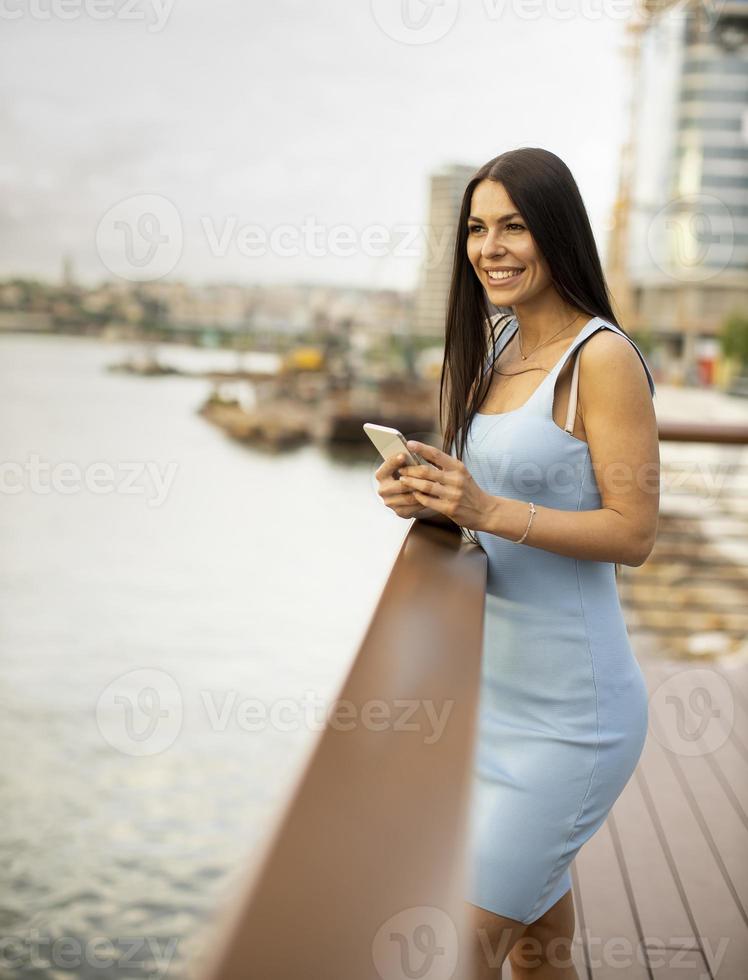 junge Frau, die ein Mobiltelefon benutzt, während sie auf der Flusspromenade steht foto
