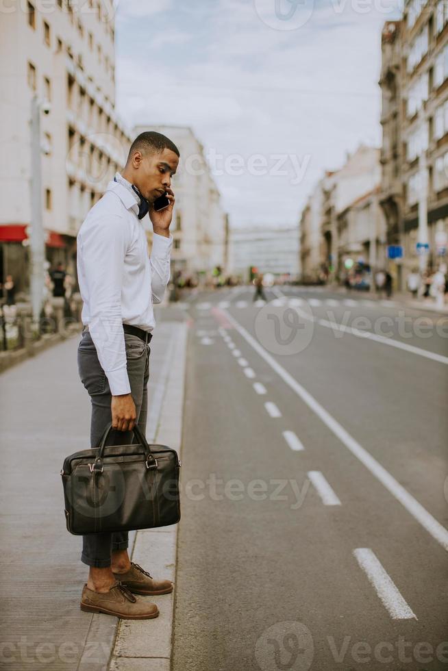 junger afroamerikanischer Geschäftsmann, der ein Mobiltelefon benutzt, während auf ein Taxi auf einer Straße wartet foto