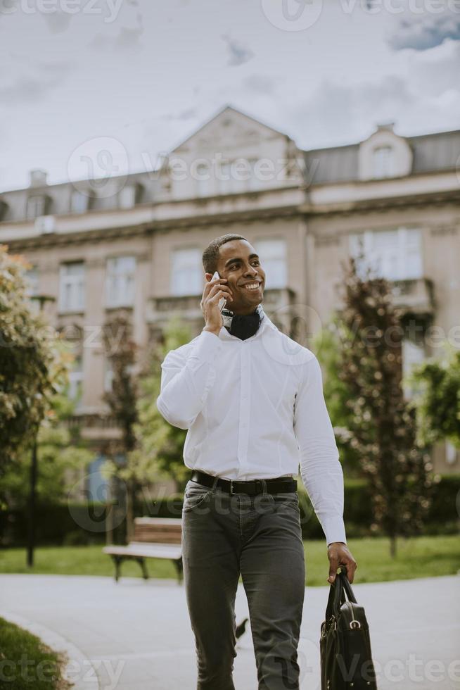 junger afroamerikanischer Geschäftsmann, der ein Mobiltelefon benutzt, während auf ein Taxi auf einer Straße wartet foto