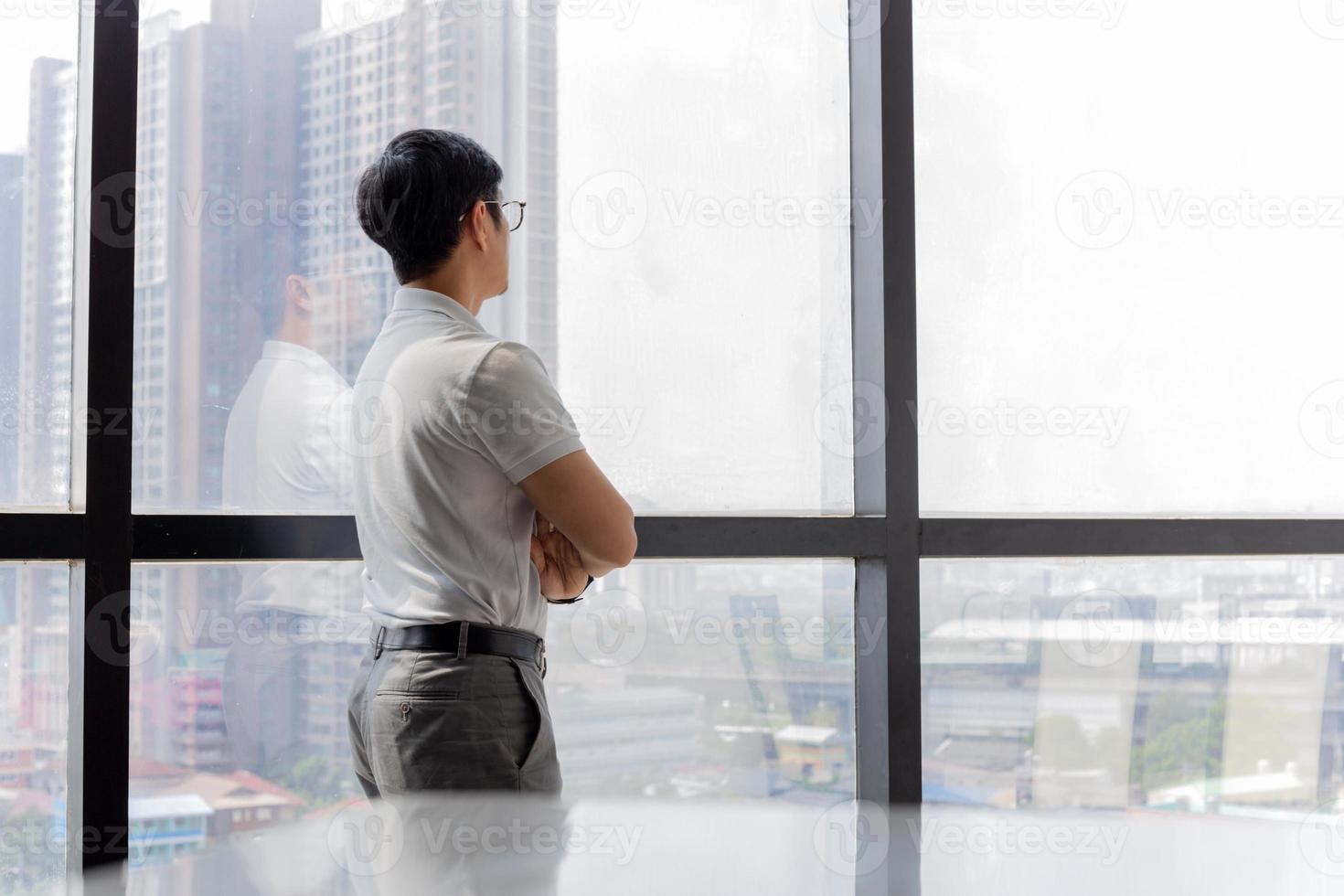 Geschäftsmann, der im Büro mit gekreuzten Händen steht und durch Fenstergebäude schaut foto