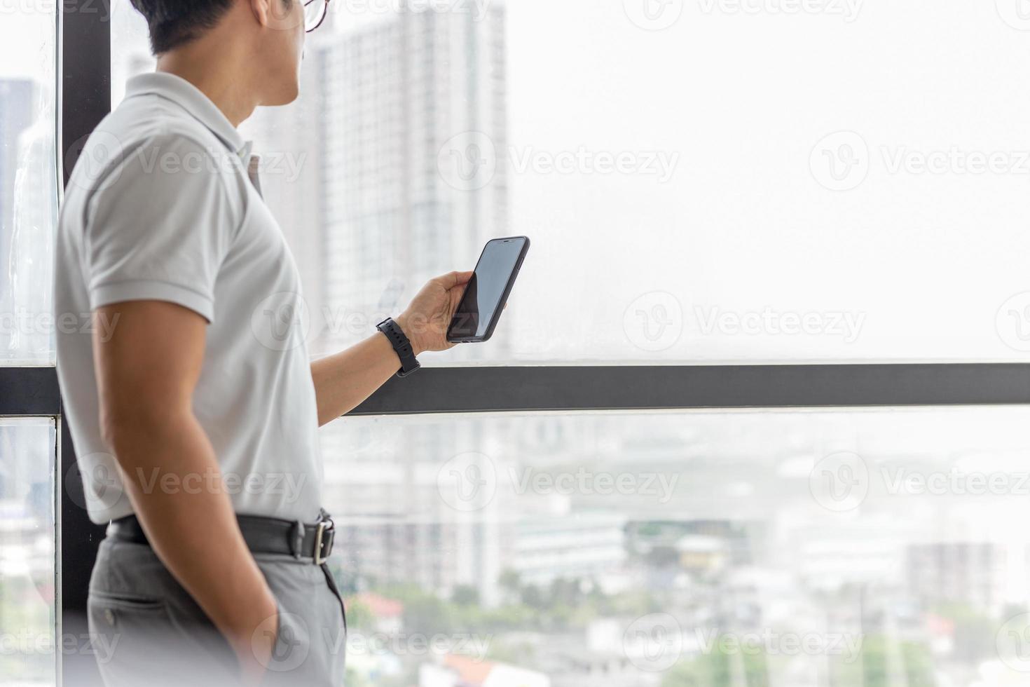 Geschäftsmann, der neben dem großen Fenster im modernen Gebäude steht, das Handy sucht foto