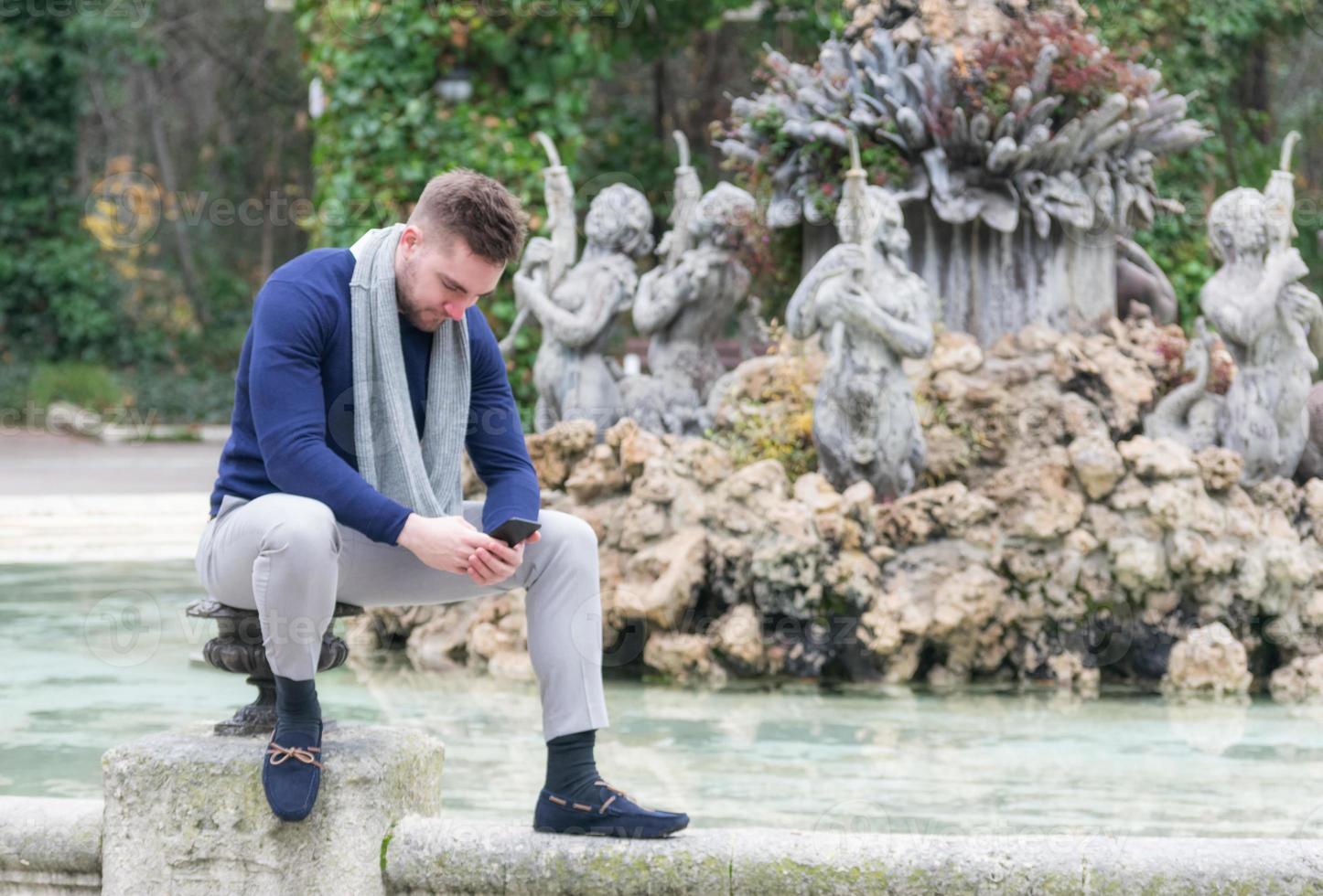 junger Mann, der im Brunnen eines Parks sitzt und sein Handy betrachtet foto