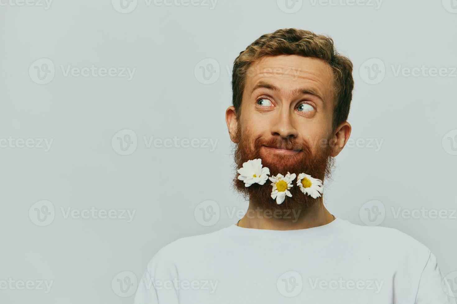 Porträt von ein komisch Mann im ein Weiß T-Shirt mit Blumen Gänseblümchen im seine Bart auf ein Weiß isoliert Hintergrund, Kopieren Ort. Urlaub Konzept und Glückwunsch. foto