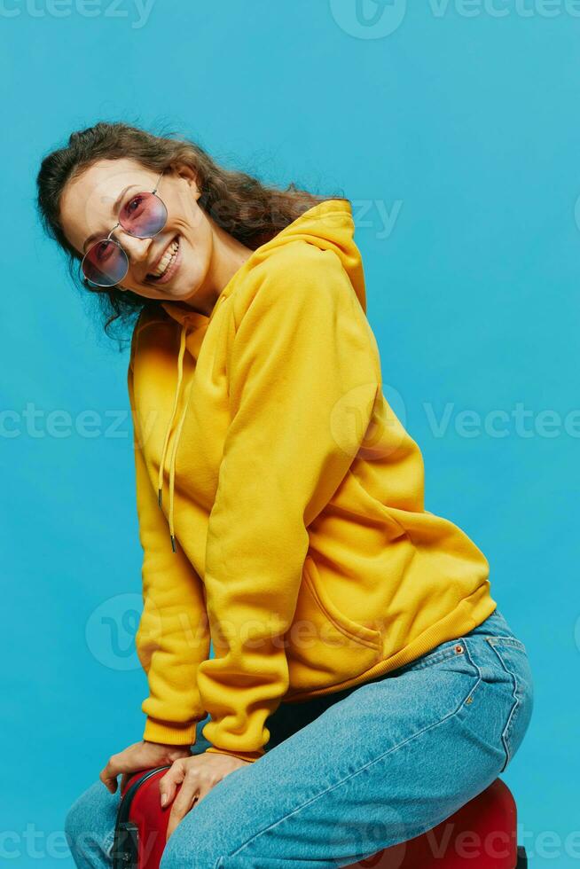 Lächeln Frau Sitzung auf ein Koffer im ein Gelb Kapuzenpullover, Blau Jeans und Brille auf ein Blau Hintergrund, Verpackung zum ein Ausflug foto