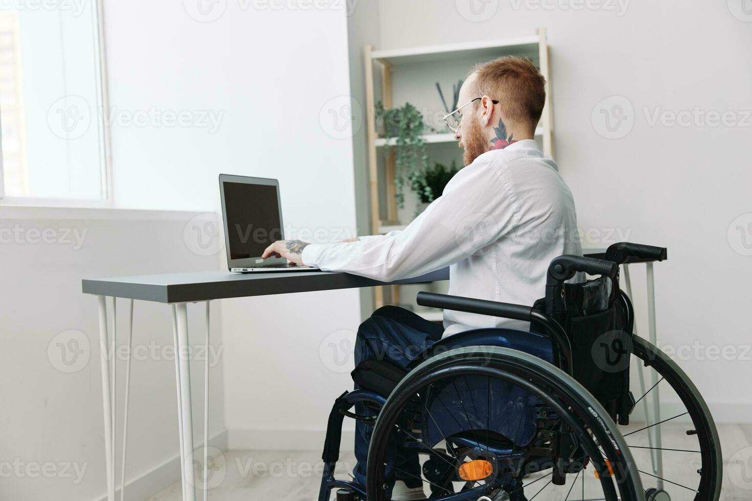 ein Mann Rollstuhl Geschäftsmann mit Tätowierungen im das Büro funktioniert beim ein Laptop online, Geschäft Verfahren, ein Rollstuhl Nahaufnahme, Integration in Gesellschaft, das Konzept von Arbeiten ein Person mit Behinderungen foto