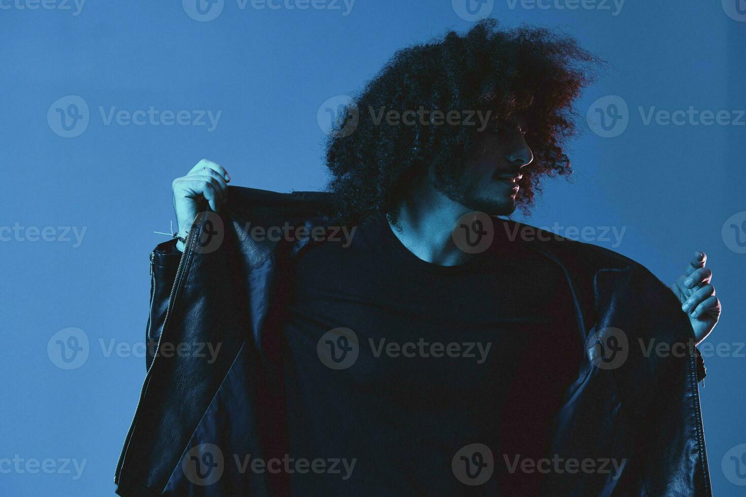 Porträt von Mode Mann mit lockig Haar auf Blau Hintergrund multinational, farbig Licht, schwarz Leder Jacke Trend, modern Konzept. foto