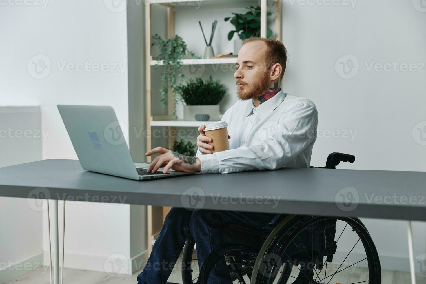 ein Mann im ein Rollstuhl ein Geschäftsmann mit Tätowierungen im das Büro funktioniert beim ein Laptop mit Kaffee, Integration in Gesellschaft, das Konzept von Arbeiten ein Person mit Behinderungen, Freiheit von Sozial Rahmen foto