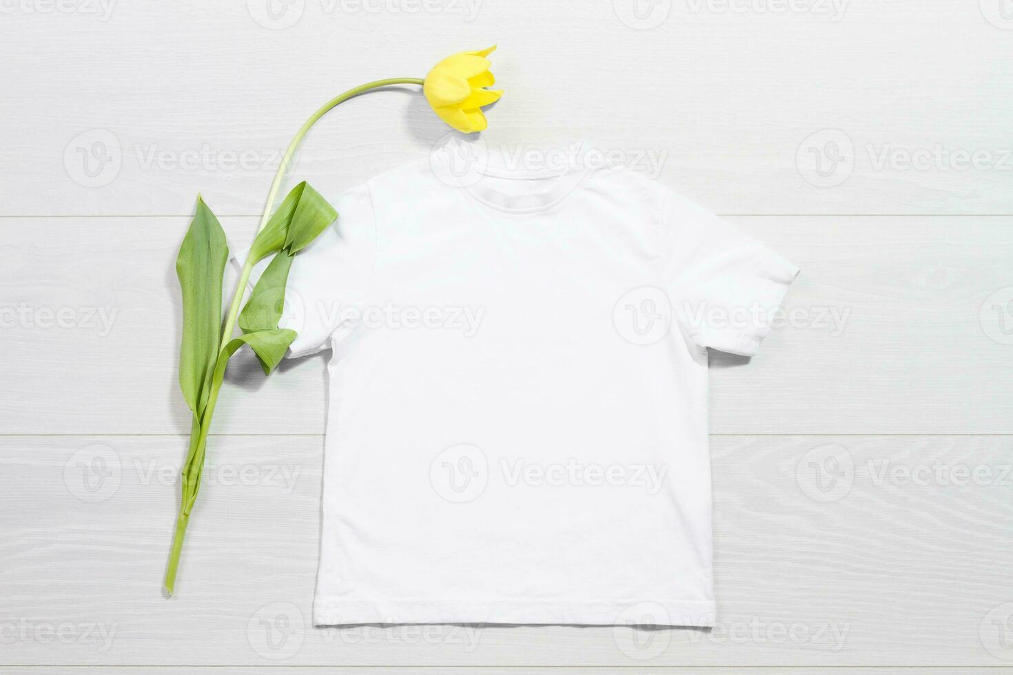 Weiß Kinder T-Shirt Attrappe, Lehrmodell, Simulation. Vorlage leer Kinder Hemd oben Sicht. Weiß hölzern Hintergrund. Mutter Frauen Tag Urlaub. Gelb Tulpe. Baby T-Shirt mit Geburtstag Blume. Frühling sehen. foto