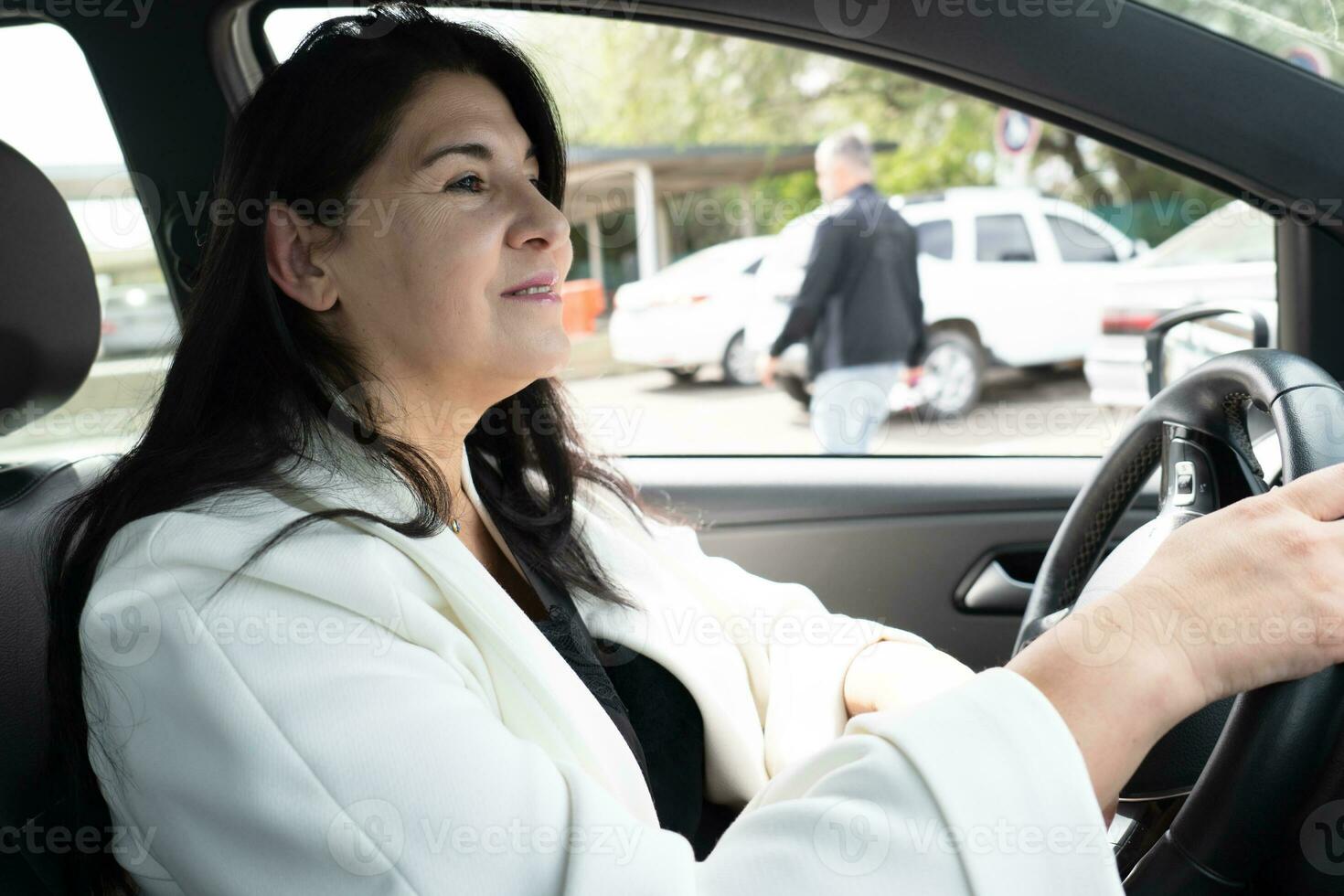 Seite Porträt von reifen Geschäft Frau im elegant Kleidung, Sitzung im modern Auto im Fahrer Sitz, lächelnd suchen aufmerksam während Fahren. Geschäft, erfolgreich Menschen, Transport Konzept. foto