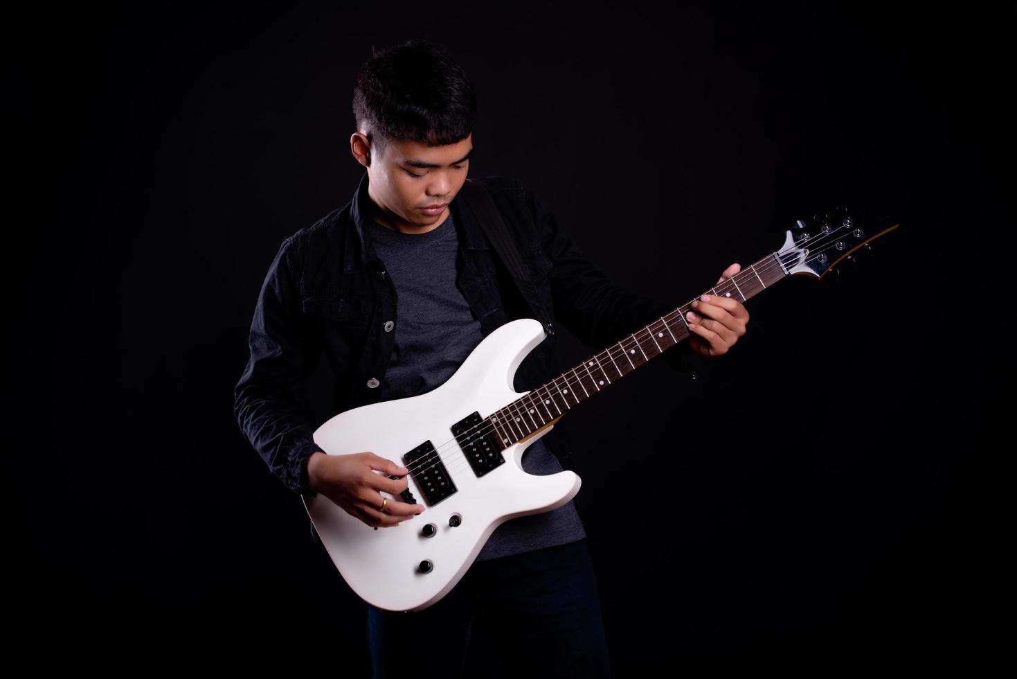 junger Mann in der schwarzen Lederjacke mit E-Gitarre gegen schwarzen Hintergrund im Studio foto