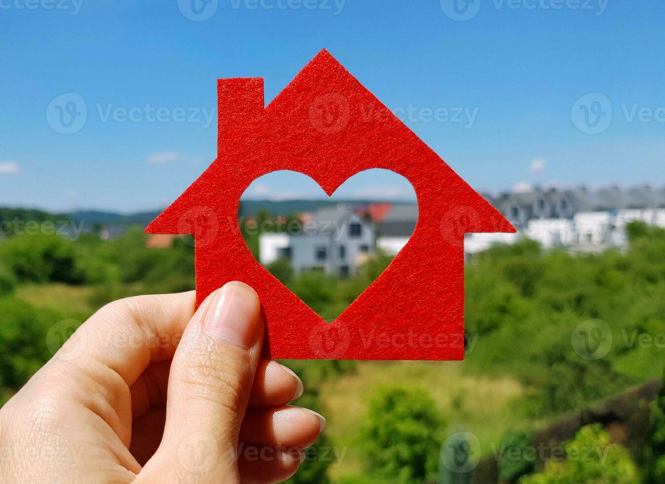 Hand halten rot fühlte Haus auf verschwommen Hintergrund von Hütte Dorf im Sommer- sonnig Tag. herzförmig Loch ist Schnitt stattdessen von ein Fenster. Kaufen oder bauen Ihre Traum heim, echt Nachlass Agentur Konzept. foto