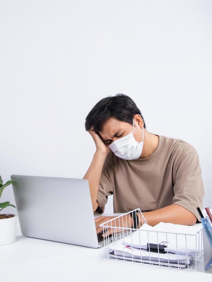 junger asiatischer mann, der eine schutzmaske trägt, fühlt sich im büro schlecht, er hat stressige kopfschmerzen von der arbeit foto
