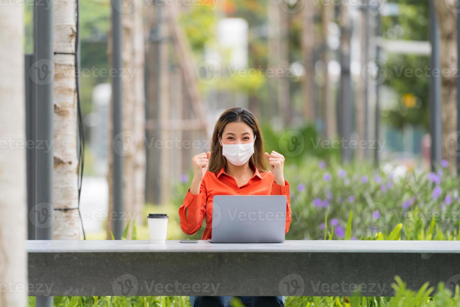 Porträt einer jungen Frau mit Gesichtsmaske, die einen Laptop im Park trägt foto