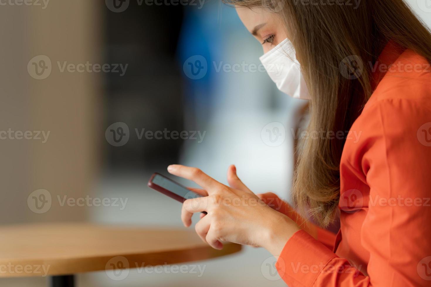 Porträt einer jungen Frau, die eine Gesichtsmaske trägt, die ein Telefon verwendet, das in der Nähe des Fensters im Café sitzt foto