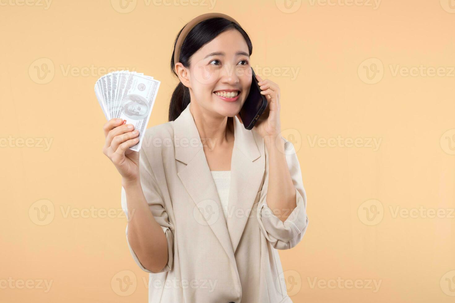 Porträt von glücklich erfolgreich zuversichtlich jung asiatisch Geschäft Frau tragen Weiß Jacke halten Smartphone und Kasse Geld Dollar Stehen Über Beige Hintergrund. Millionär Geschäft, Einkaufen Konzept. foto