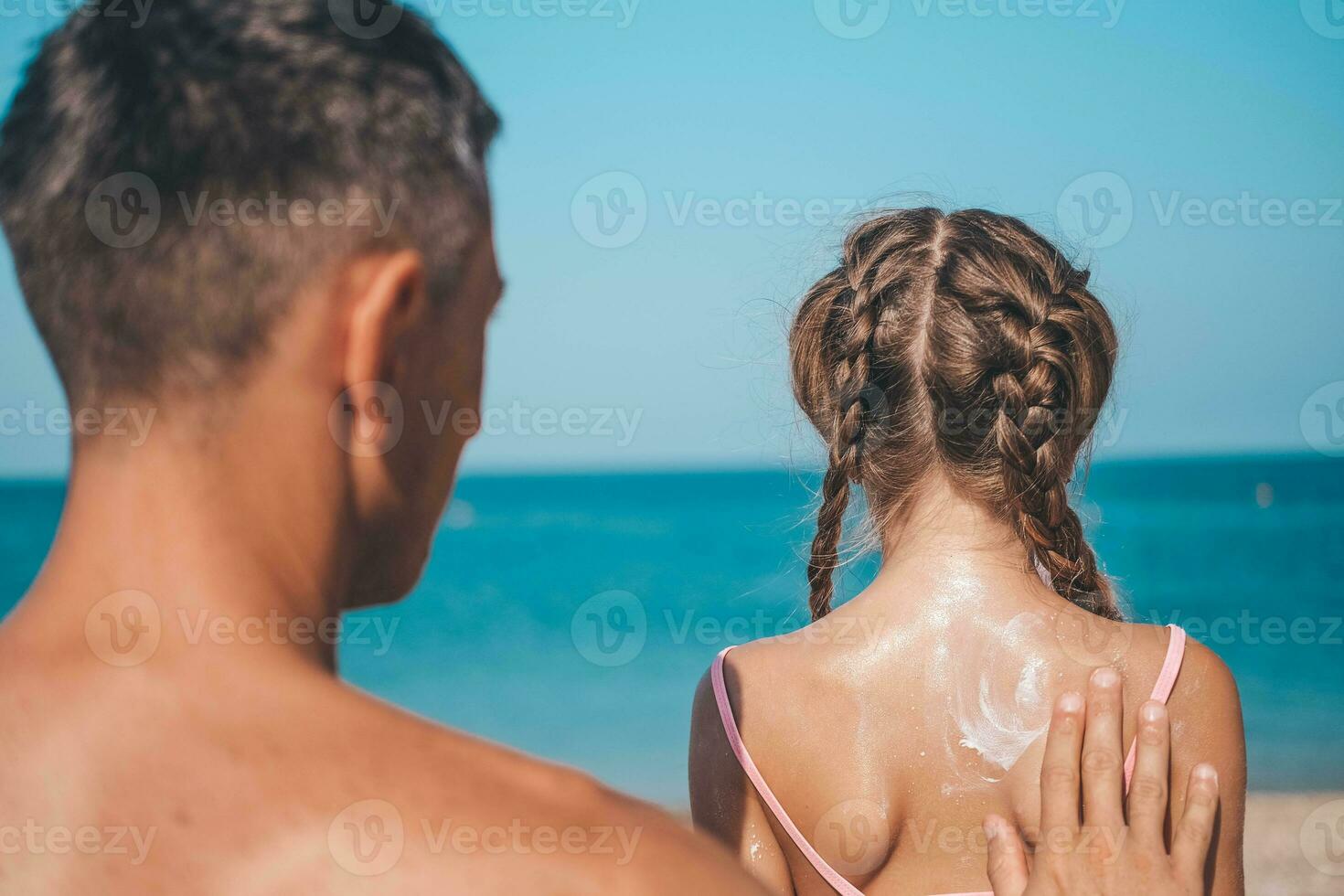 Vater bewirbt sich schützend Sahne zu ihr Tochter Gesicht beim das Strand.  Mann Hand halten Sonnenschutz Lotion auf Baby Gesicht. süß wenig Mädchen  mit ein Sonne Block durch das Meer . Kopieren