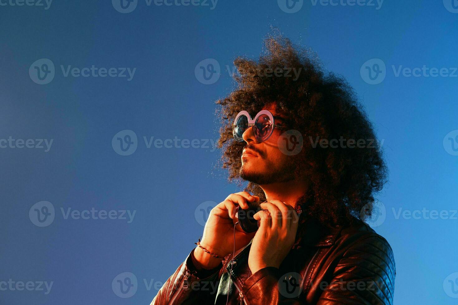 Porträt von ein stilvoll Mann mit lockig Haar mit Brille und Kopfhörer auf ein Blau Hintergrund multinational, farbig Licht, schwarz Leder Jacke Trend, modern Konzept. foto