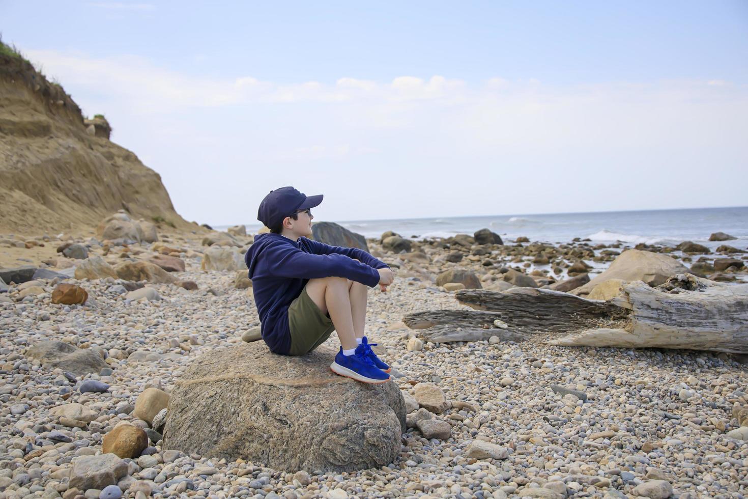 Junge sitzt auf einem Felsen am Strand und schaut auf das Meer foto