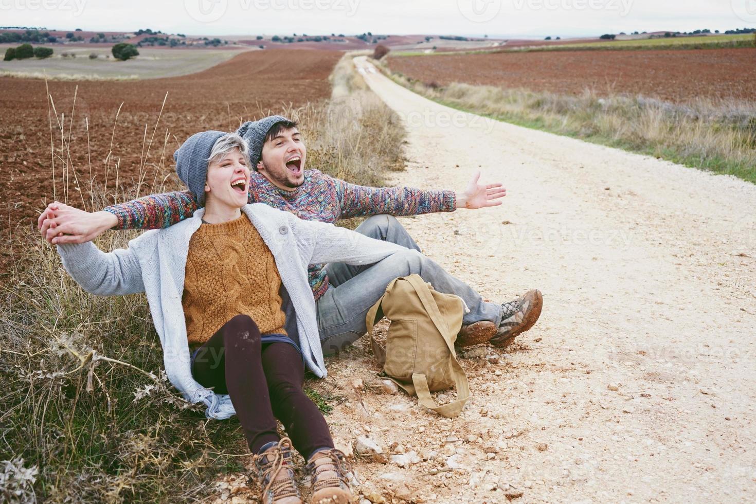 Ein paar junge Millennials, die sich bei einer Abenteuerreise auf einem Feldweg im Freien umarmen und an einem Rucksack schreien foto