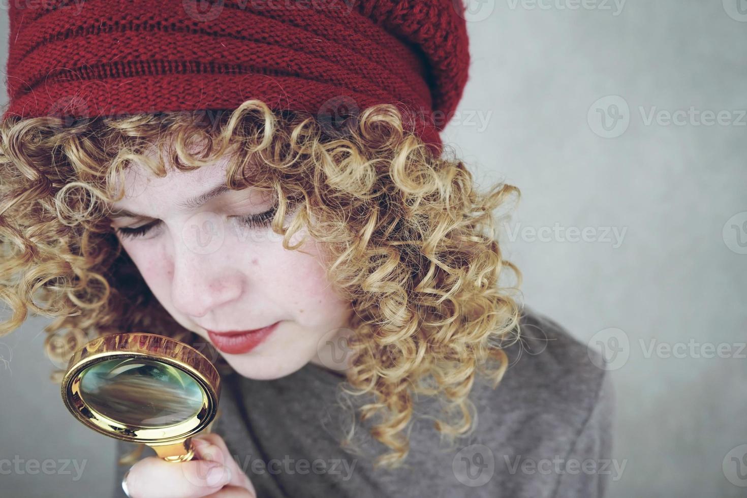 Nahaufnahmeporträt einer schönen und jungen lustigen Frau mit blauen Augen und lockigen blonden Haaren, die mit einer Lupe untersucht foto