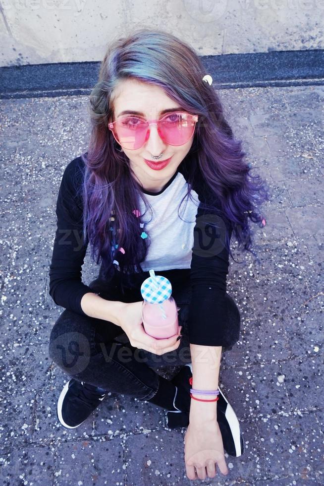 glücklicher schöner Teenager mit rosa Sonnenbrille trinkt und genießt ein rosa Getränk, das auf städtischen Boden sitzt foto