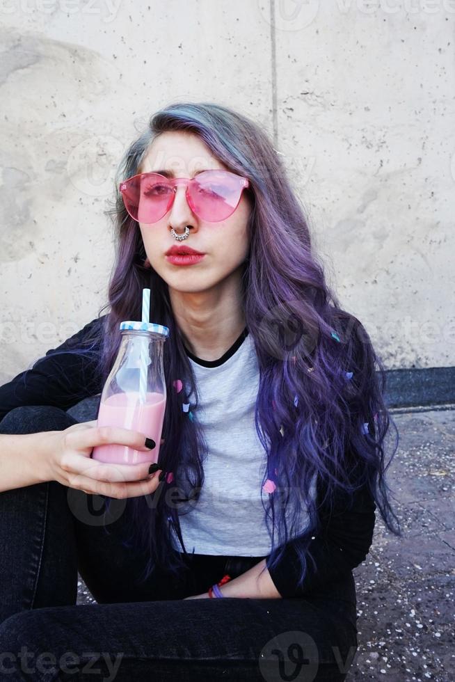 glücklicher schöner Teenager mit rosa Sonnenbrille trinkt und genießt ein rosa Getränk, das auf städtischen Boden sitzt foto