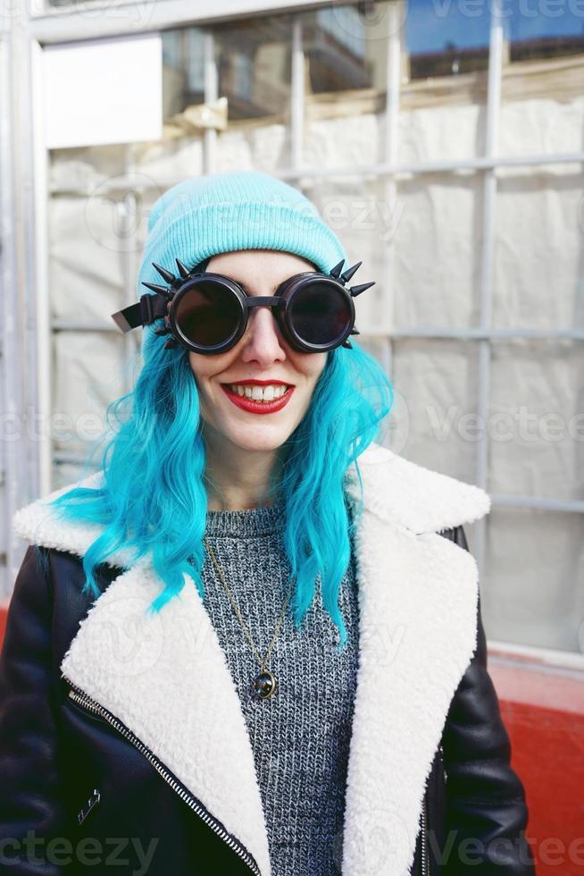 Porträt einer jungen Punk- oder Gothic-Frau lächelt mit blau gefärbten Haaren und trägt eine schwarze Steampunk-Brille und eine blaue Wollmütze in einer städtischen Straße im Freien foto