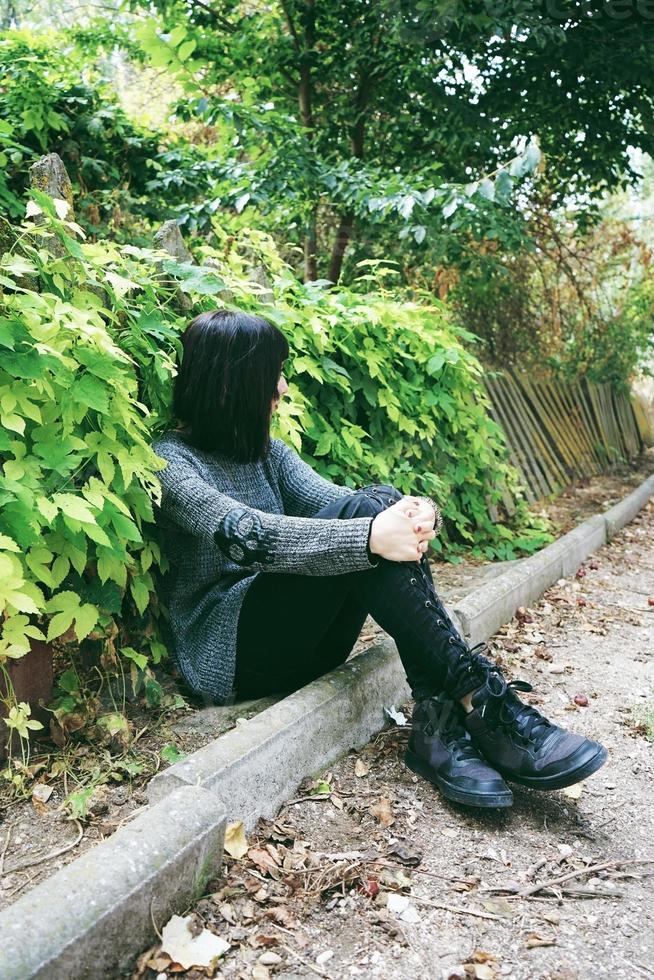 Schöne kaukasische brünette Frau, die auf dem Boden in einem Park sitzt und Punk- oder Gothic-Kleidung trägt und von grünen Blättern umgeben ist foto