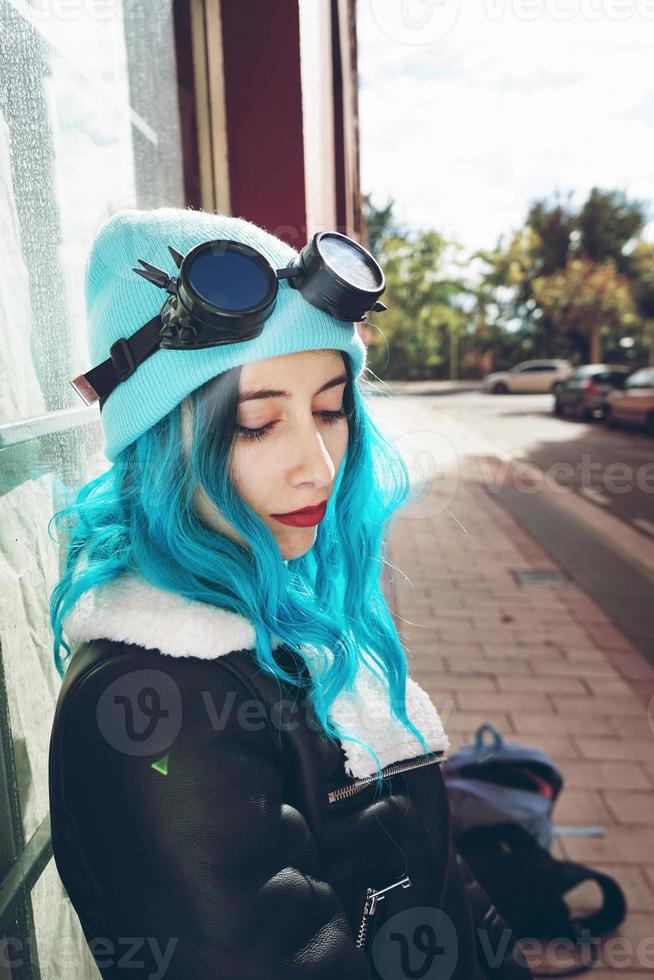 Porträt einer jungen Punk- oder Gothic-Frau mit blauen Haaren und schwarzer Steampunk-Brille und blauer Wollmütze in einer städtischen Straße im Freien foto