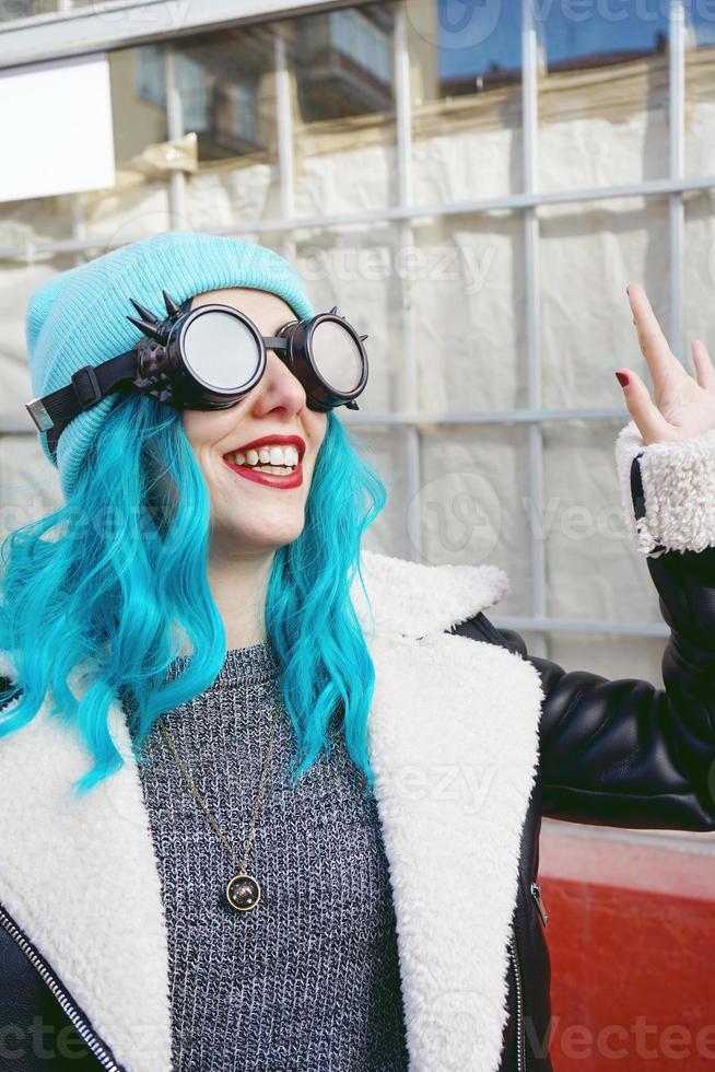 Porträt einer jungen Punk- oder Gothic-Frau lächelt mit blau gefärbten Haaren und trägt eine schwarze Steampunk-Brille und eine blaue Wollmütze in einer städtischen Straße im Freien foto