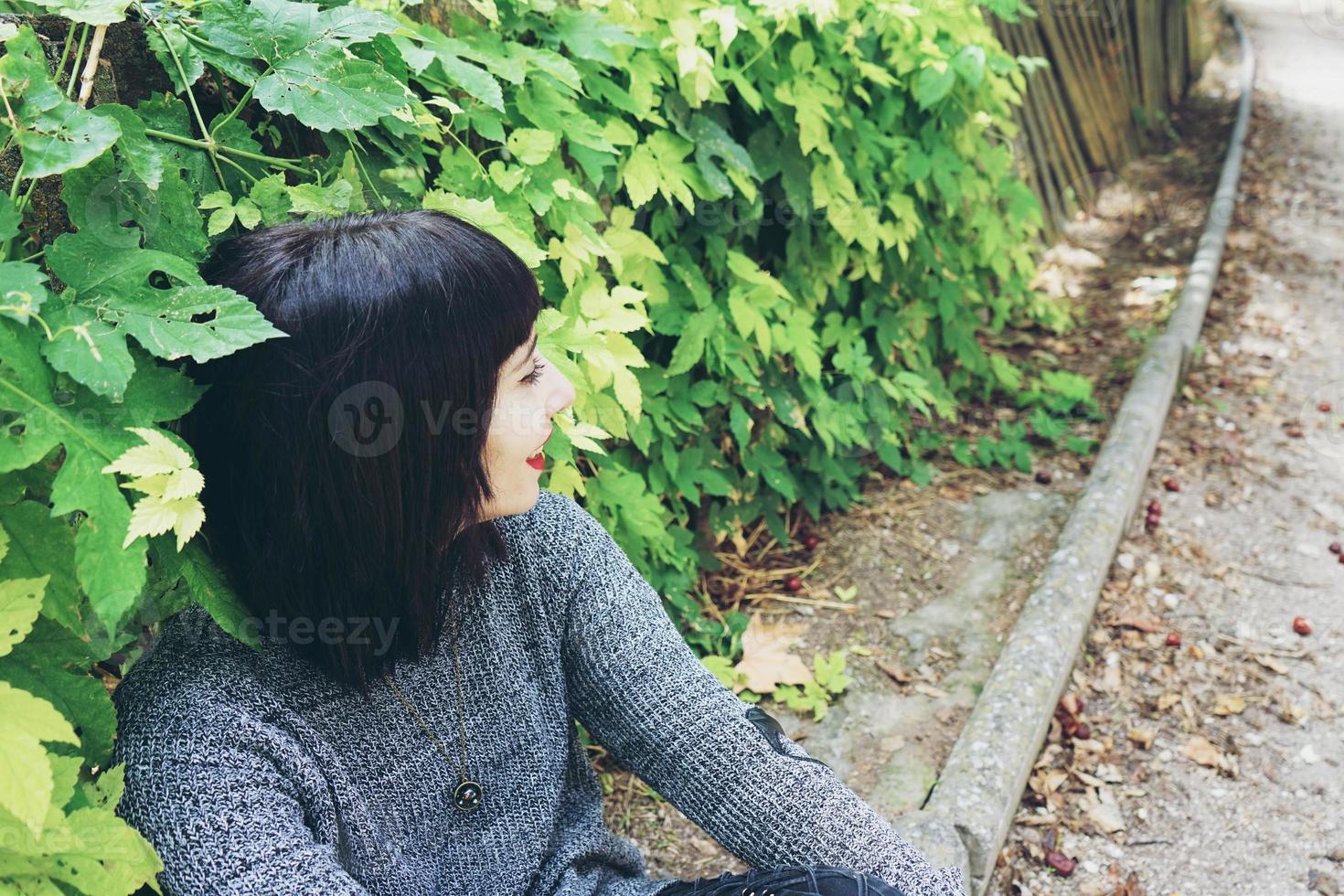 Schöne kaukasische brünette Frau, die auf dem Boden in einem Park sitzt und Punk- oder Gothic-Kleidung trägt und von grünen Blättern umgeben ist foto