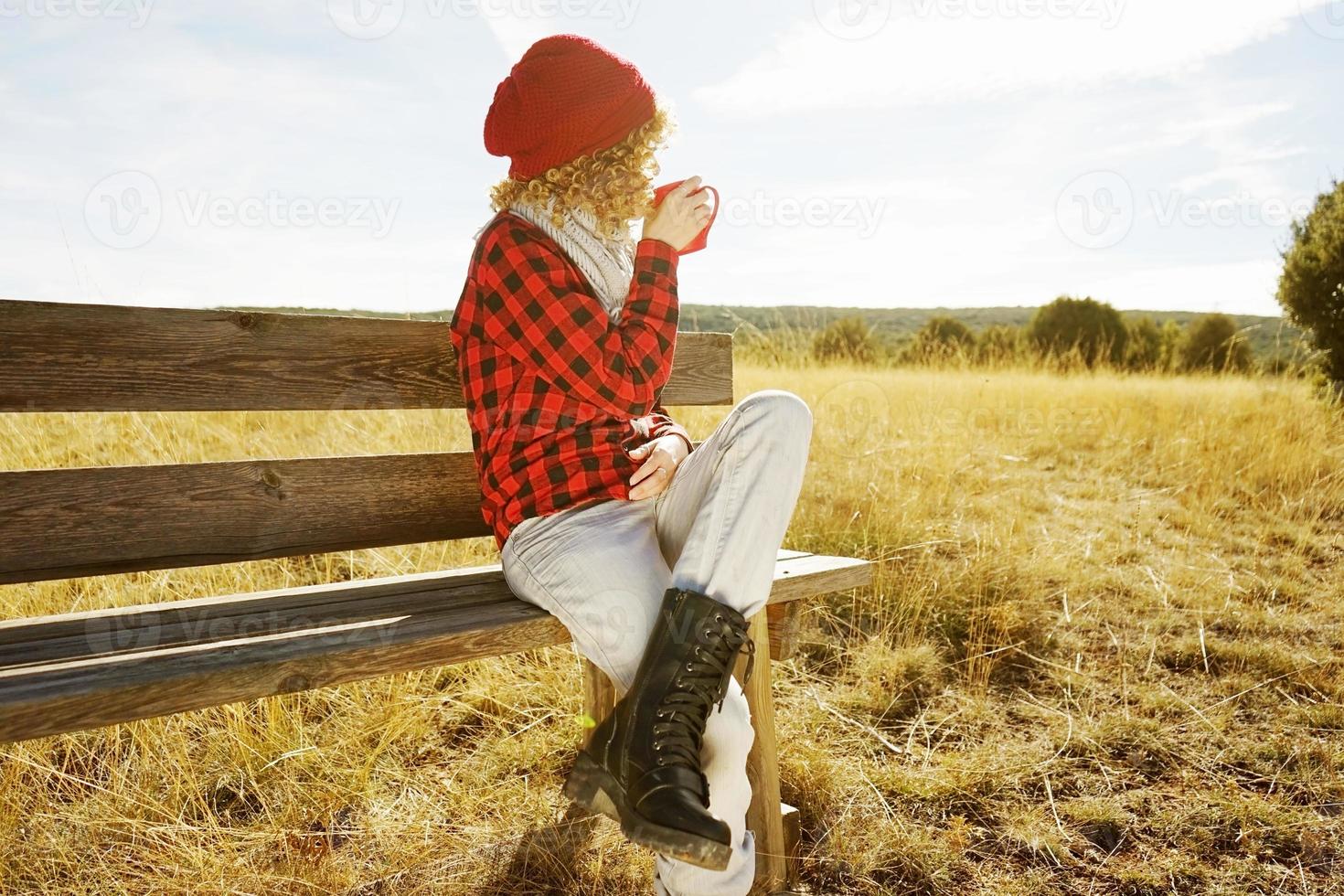 eine junge Frau von hinten in rotem kariertem Hemd mit Wollmütze und Schal, die eine Tasse Tee oder Kaffee trinkt, während sie auf einer Holzbank in einem gelben Feld mit Hintergrundbeleuchtung von der Herbstsonne sitzt foto