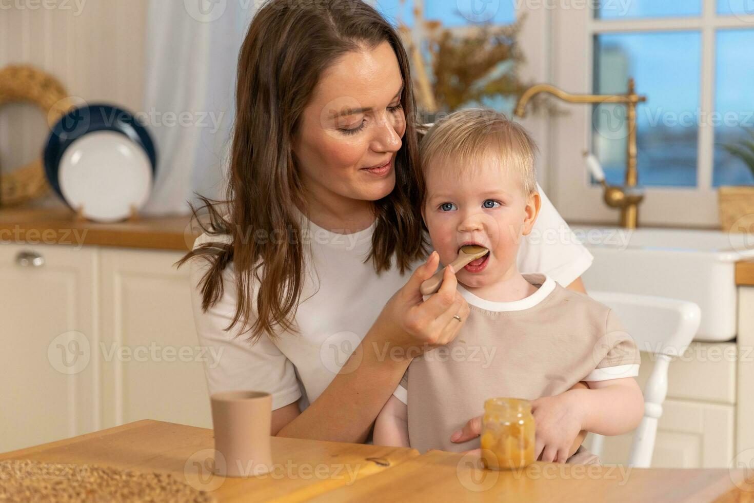 glücklich Familie beim heim. Mutter Fütterung ihr Baby Junge von Löffel im Küche. wenig Kleinkind Kind mit unordentlich komisch Gesicht isst gesund Essen beim heim. jung Frau Mama geben Essen zu Kind Sohn. foto