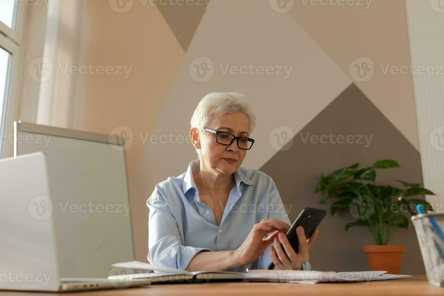 zuversichtlich stilvoll europäisch Mitte alt Senior Frau mit Smartphone beim Arbeitsplatz. stilvoll älter reifen 60er Jahre grau behaart Dame Geschäftsfrau mit Zelle Telefon im Büro. Boss Führer mit Internet Anwendungen. foto