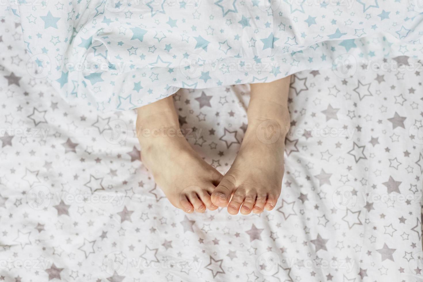 Nahaufnahme der Füße des Kleinkindmädchens auf dem Bett unter der Decke. helle Blau- und Beigetöne. foto