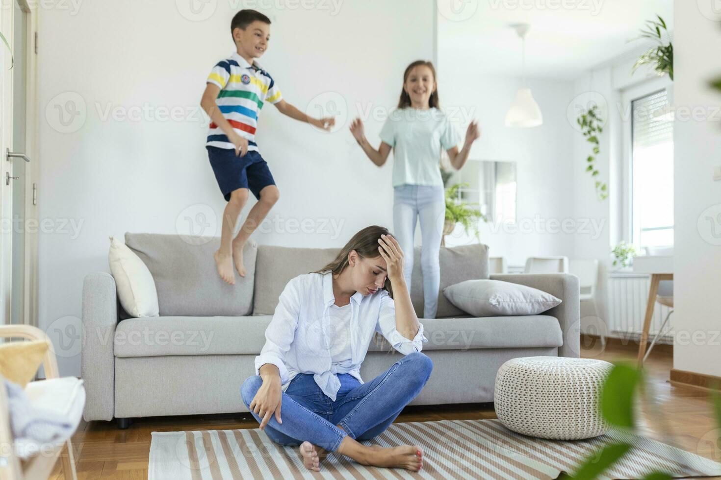 müde jung Mutter Sitzung auf das Fußboden und Arbeiten mit Laptop und Unterlagen während wenig Kinder und Springen auf das Sofa und haben Spaß und Herstellung Lärm foto