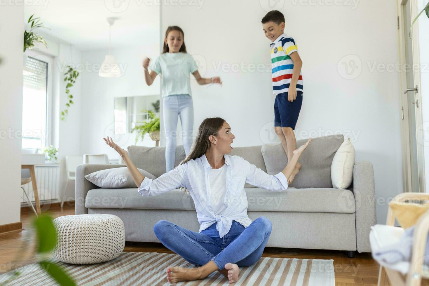 müde jung Mutter Sitzung auf das Fußboden und Arbeiten mit Laptop und Unterlagen während wenig Kinder und Springen auf das Sofa und haben Spaß und Herstellung Lärm foto