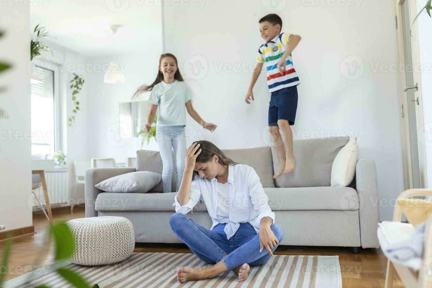 müde jung Mutter Sitzung auf das Fußboden und Arbeiten mit Laptop und Unterlagen während wenig Kinder und Springen auf das Sofa und haben Spaß und Herstellung Lärm foto