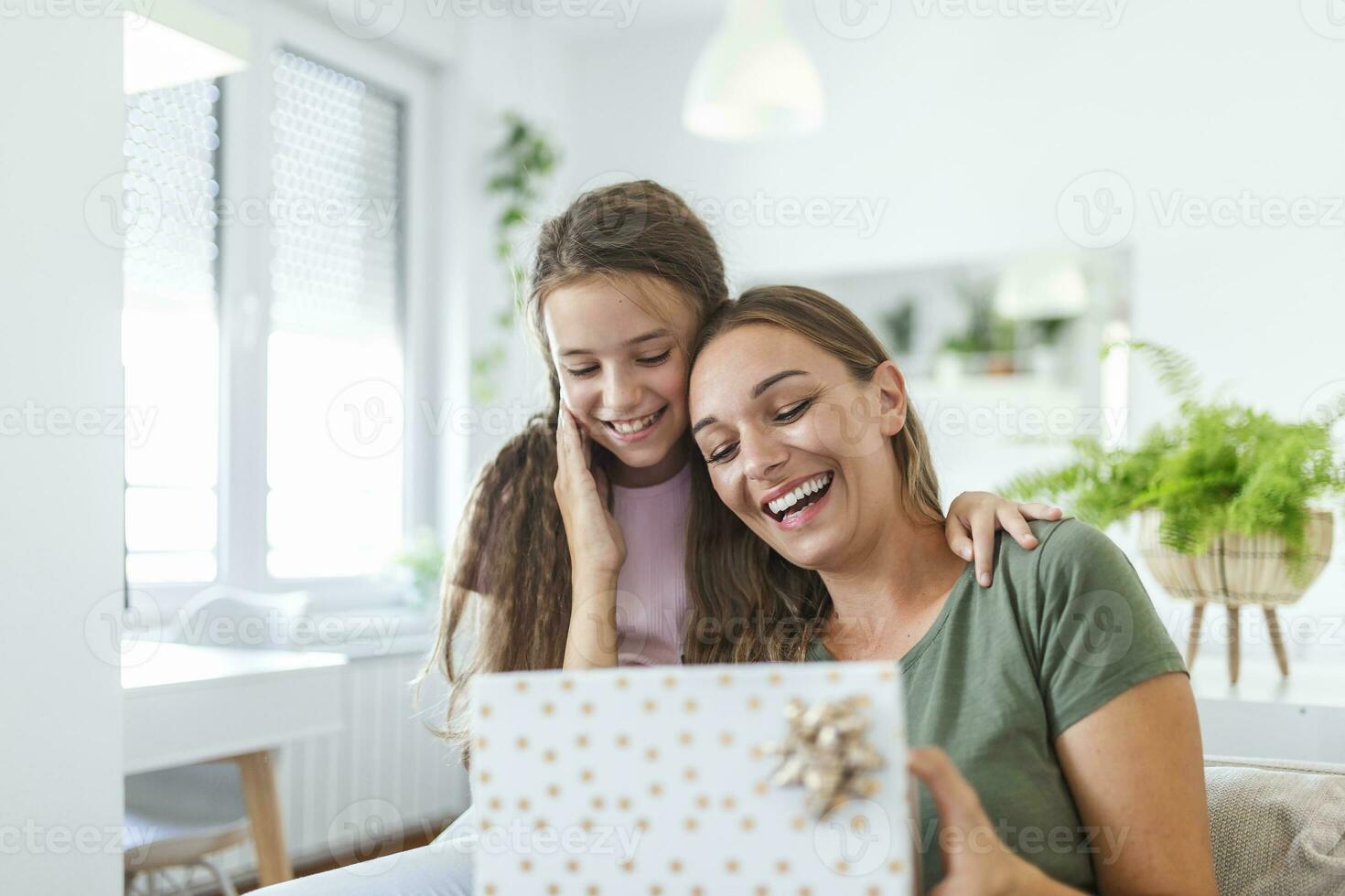 jung Frau und Mädchen beim Zuhause feiern Mutter Tag Sitzung auf Sofa Tochter umarmen Mutter küssen Wange Mama Lachen froh halten Geschenk Box foto