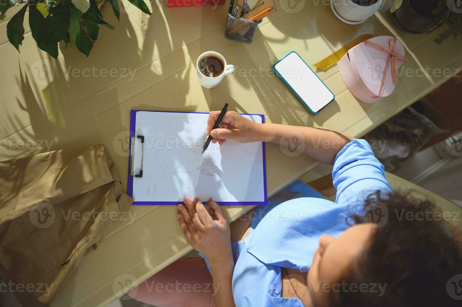 oben Sicht. jung Frau zeichnet auf Zwischenablage, Sitzung beim Schreibtisch mit ein Tasse von Kaffee, Smartphone mit Weiß Attrappe, Lehrmodell, Simulation Bildschirm foto