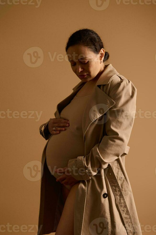 schön schwanger Frau im Beige Graben Mantel, streicheln ihr groß Bauch, Gefühl Baby Tritte. Mutterschaft. Schwangerschaft Mode foto
