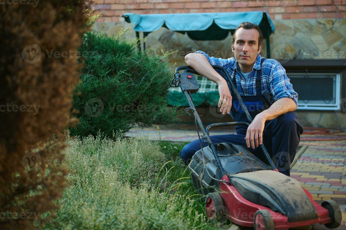 Fachmann männlich Gärtner im Gartenarbeit Uniform, Sitzung in der Nähe von elektrisch Rasen Mäher, lächelnd suchen selbstbewusst beim Kamera foto