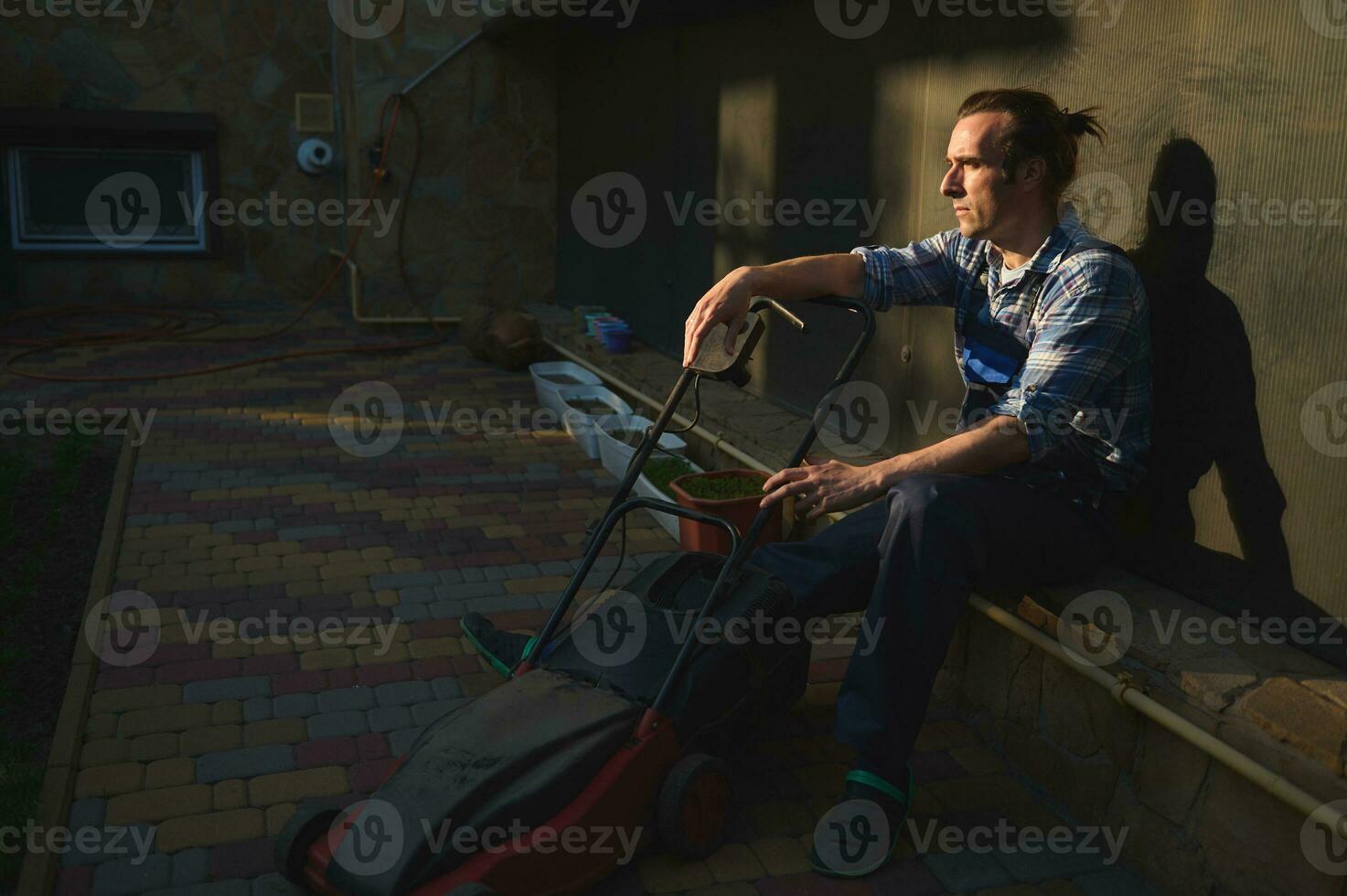 Porträt von ein müde überarbeitet Gärtner Mann mit elektrisch Rasen Mäher, entspannend nach schwer Arbeiten Tag im das Hinterhof foto