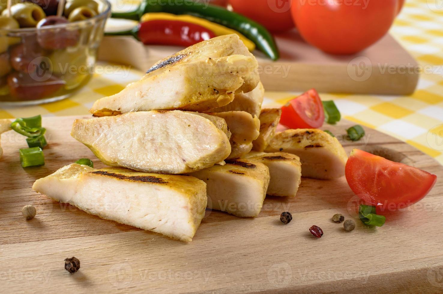 Gegrilltes Hühnerfleisch auf Holzplatte mit Gemüse und Oliven im Hintergrund. foto