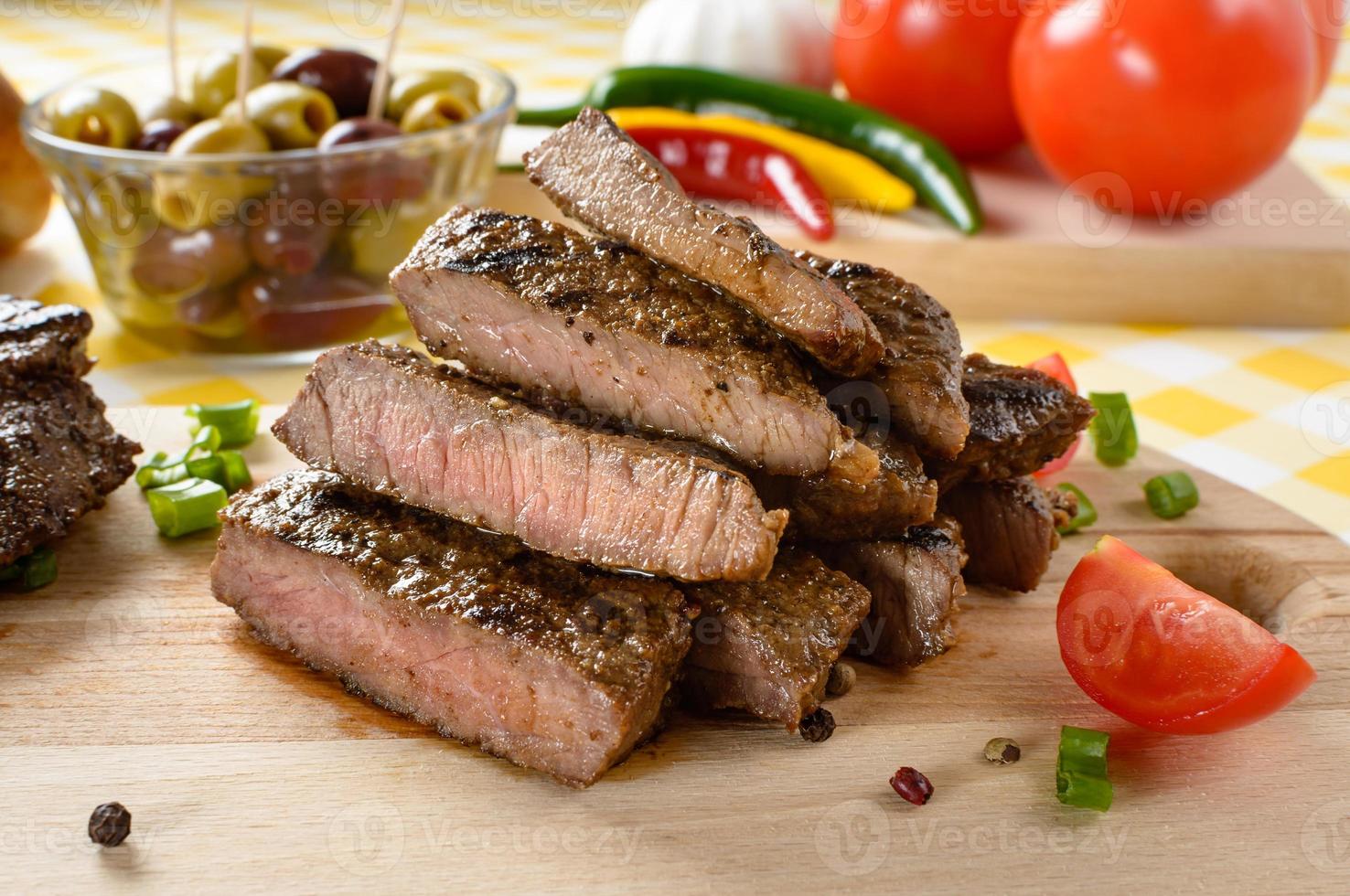 Gegrilltes Rindfleisch auf Holzplatte mit Gemüse und Oliven im Hintergrund. foto