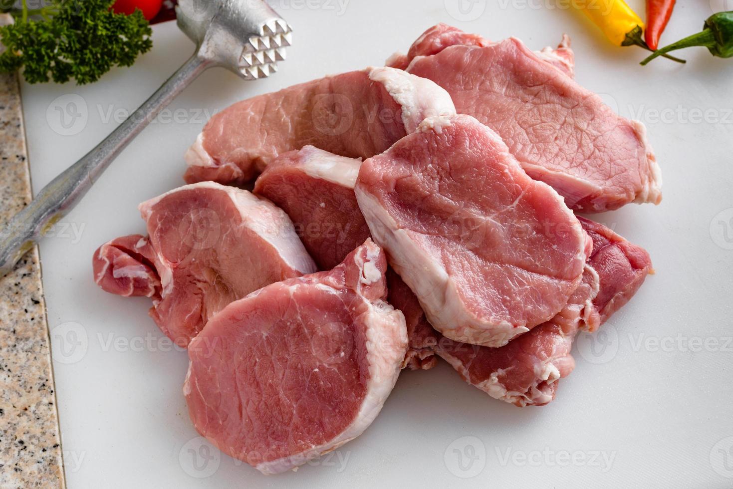 rohes Fleisch, Schweinesteaks auf dem Küchentisch. Gemüse und Fleischhammer im Hintergrund. foto