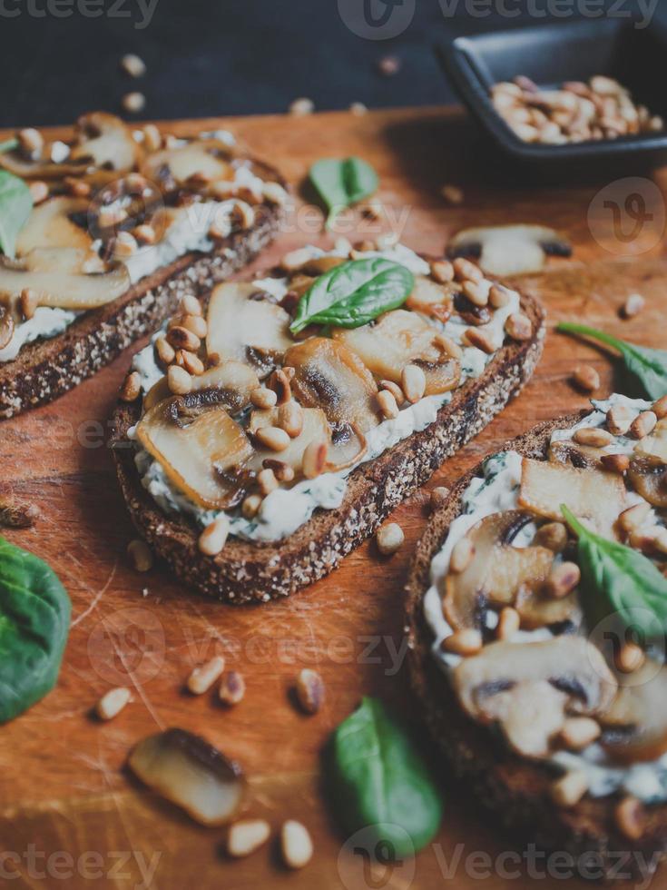 leckere frische Bruschetta mit Pilzen, Spinat, Knoblauch, Frischkäse und Pinienkernen, auf einem Holzbrett, auf dunklem Hintergrund. foto